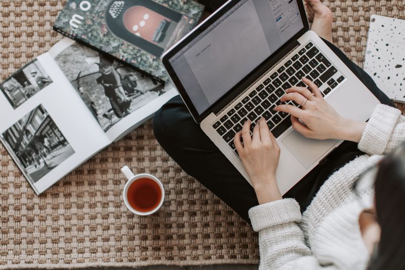 A woman typing a content on her laptop with magazine and coffee on the side