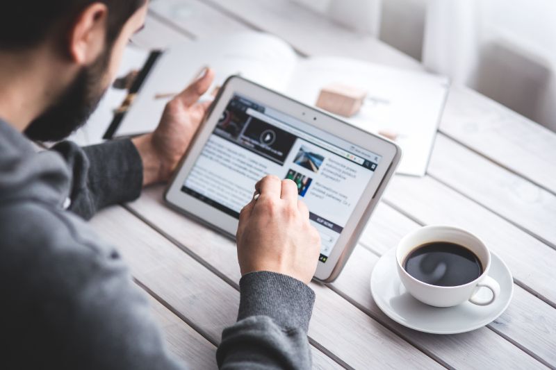 A guy holding a tablet and reading an article with a coffee on the side