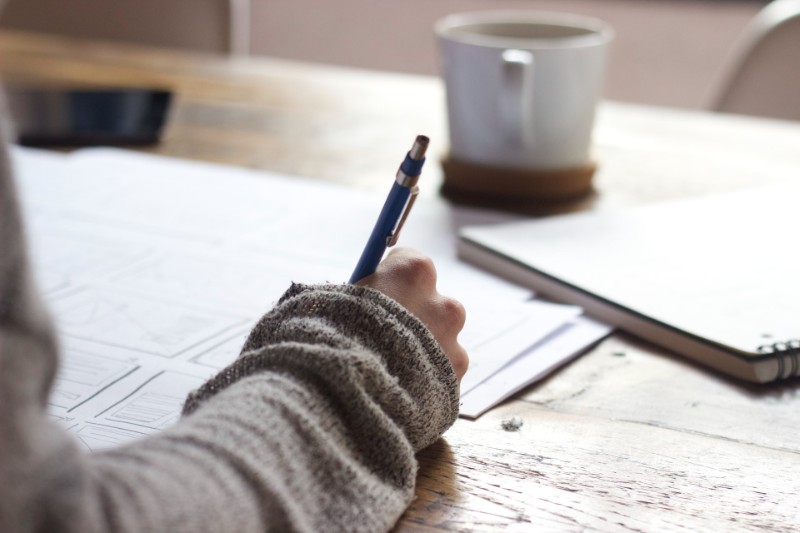 Woman wearing gray sweater writing, scattered paper, cup of coffee