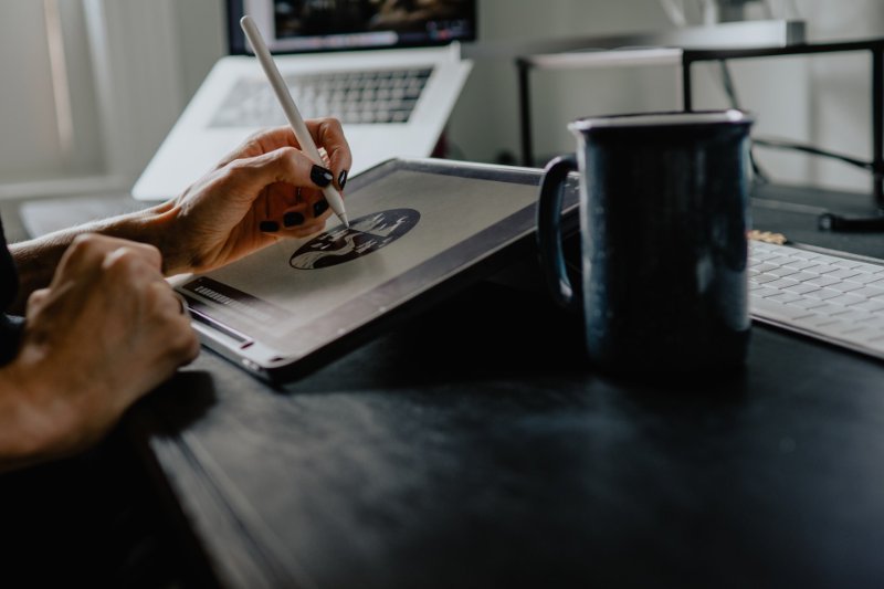 someone drawing on their device with a coffee mug and laptop on the table