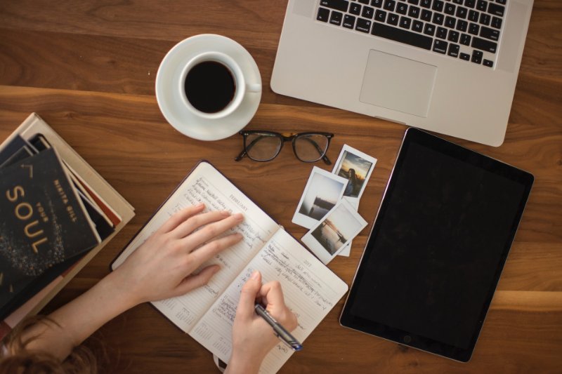 a person writing a diary with eyewear, photographs, ipad, laptop, coffee mug and books on the desk