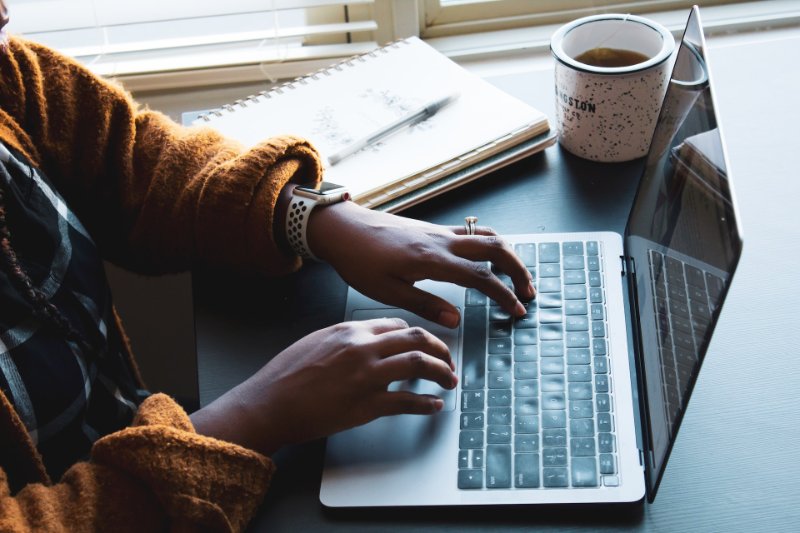 Someone typing something on the computer with tea, pen and notebook on the table