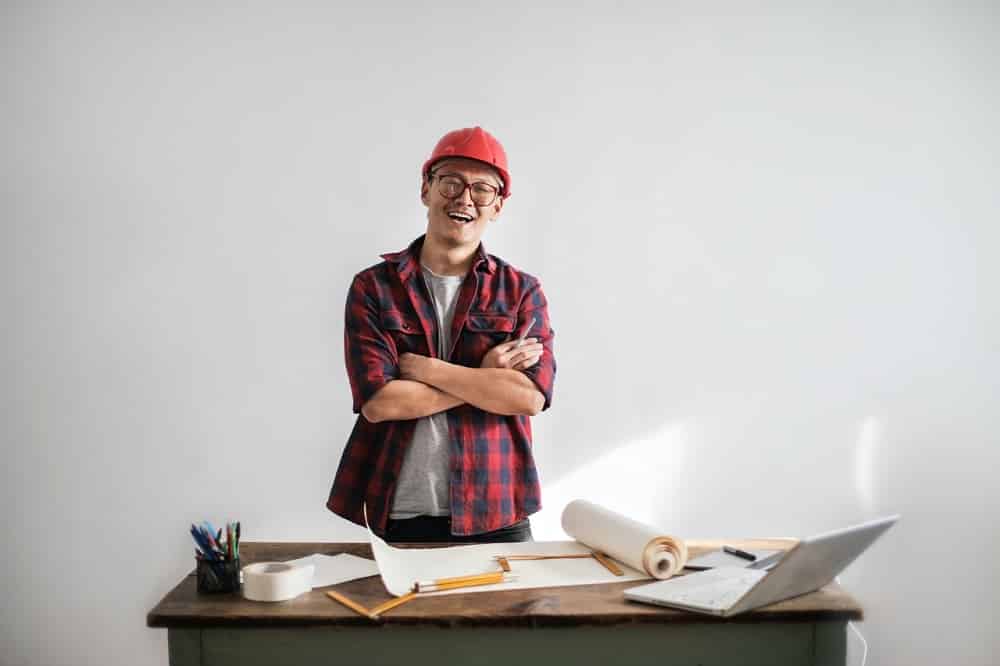 engineering expert standing with folded hands behind the table scattered with his work assignments and laptop