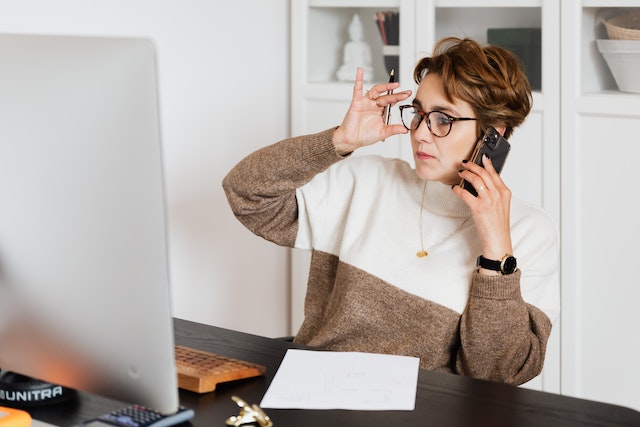 Lady with glasses working on her computer with a pen and phone in hands