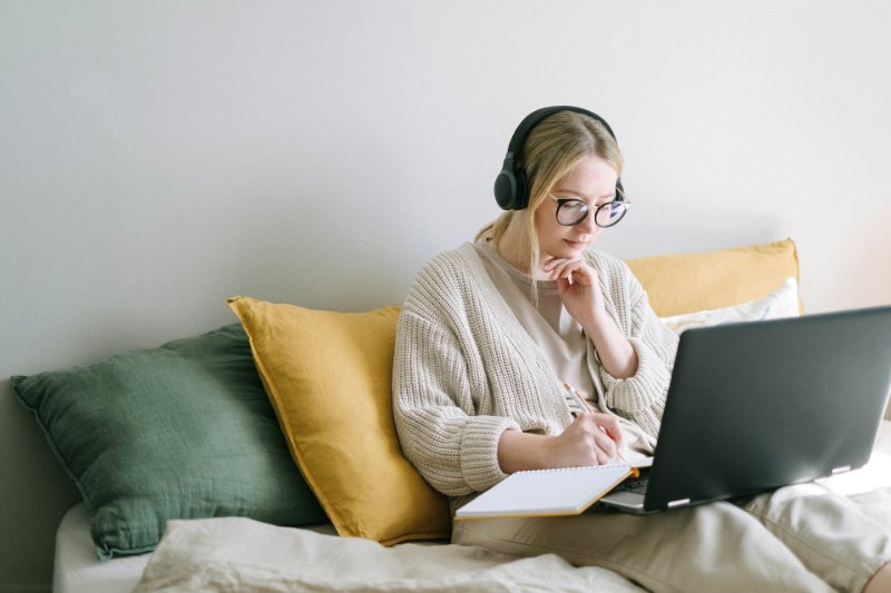 A girl wearing headphones using her laptop and taking notes