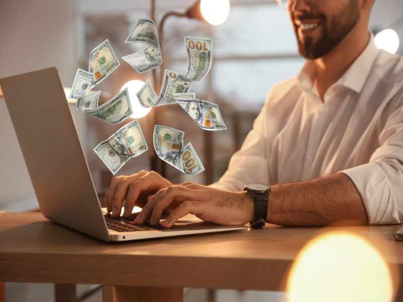 Closeup view of man using laptop at table and flying dollars