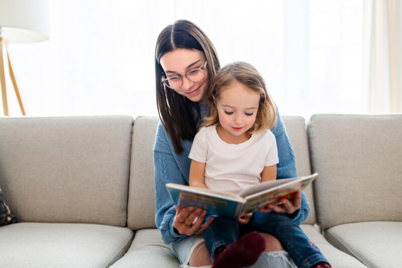 Happy kid sitting on sofa with babysitter teen holding book