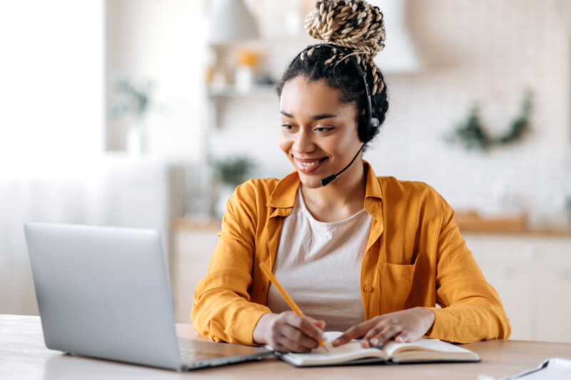 Concentrated confident friendly african american girl in headset, call center operator, manager, agent of support service, conducts online consultation, video briefing, taking notes, smiling friendly