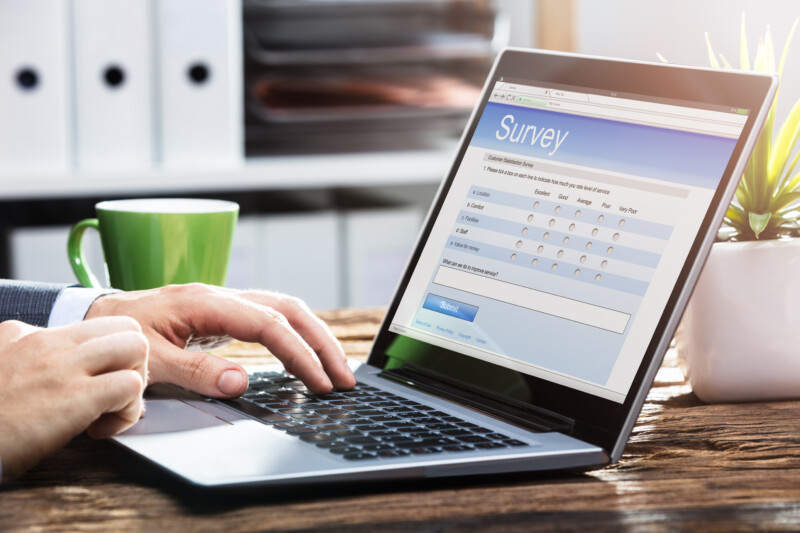 Close-up Of A Businessperson's Hand Filling Online Survey Form On Laptop Over Wooden Desk