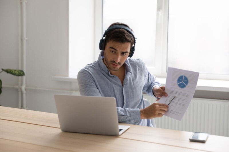 Serious marketing professional man in headphones talking to client on video call, presenting paper graphic reports, showing, explaining diagram. Employee, manager attending online meeting