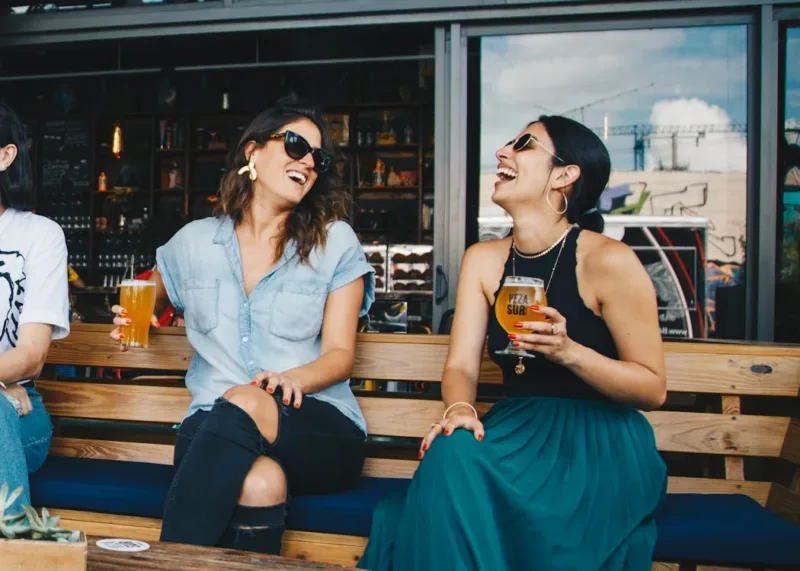 Two Smiling Women Sitting on Wooden Bench drinking
